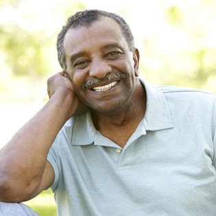 An older man wearing a polo shirt and propping his head with his hand, smiling and pleased with his full dentures