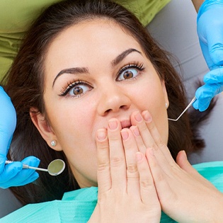 A woman lying back in a dentist’s chair with her hands clasped over her mouth, anxious about dental work