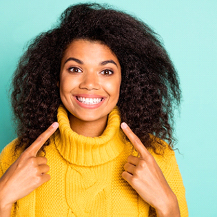 A young woman wearing a yellow sweater and pointing to her straighter, healthier smile 